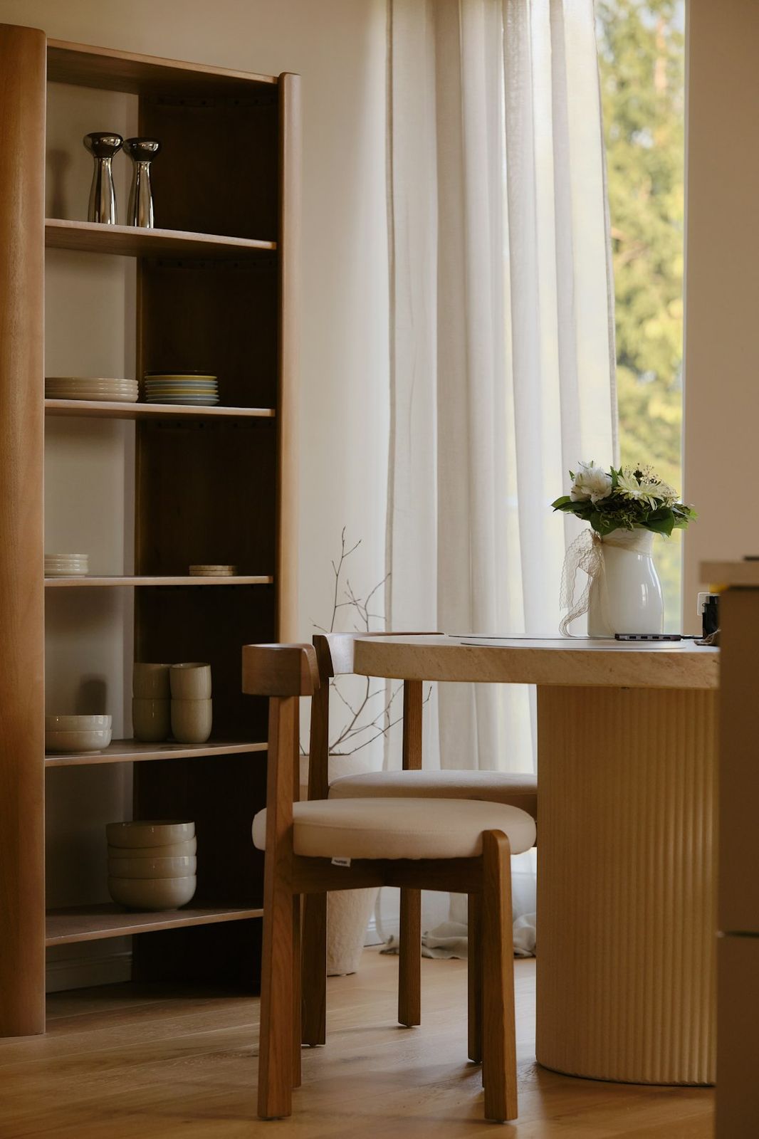 A wooden dining table with a vase of flowers beside a tall shelving unit in a sunlit room