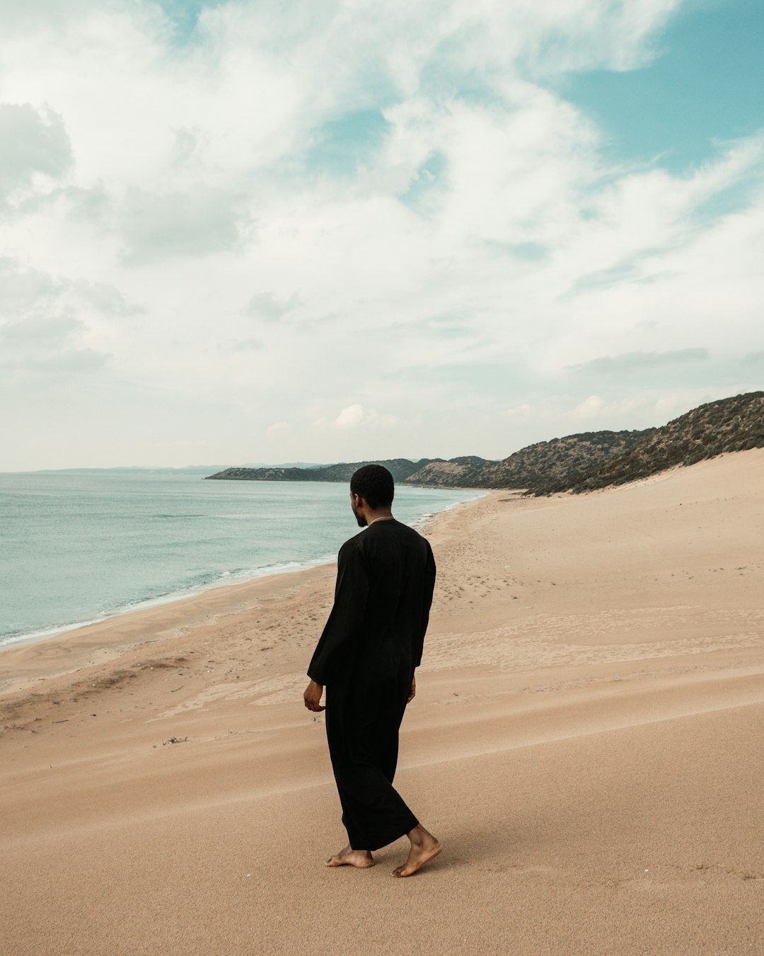 A person in a black robe walking barefoot on a sandy beach by the sea
