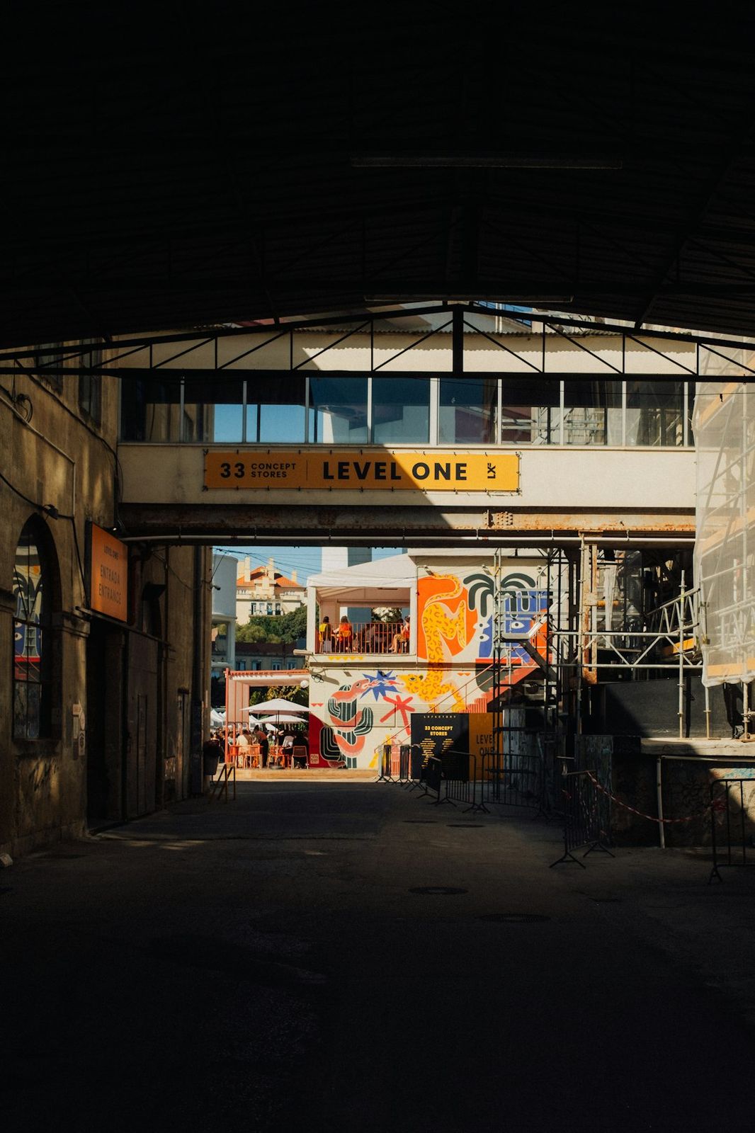 A view through a dark industrial archway toward a brightly painted building with outdoor seating