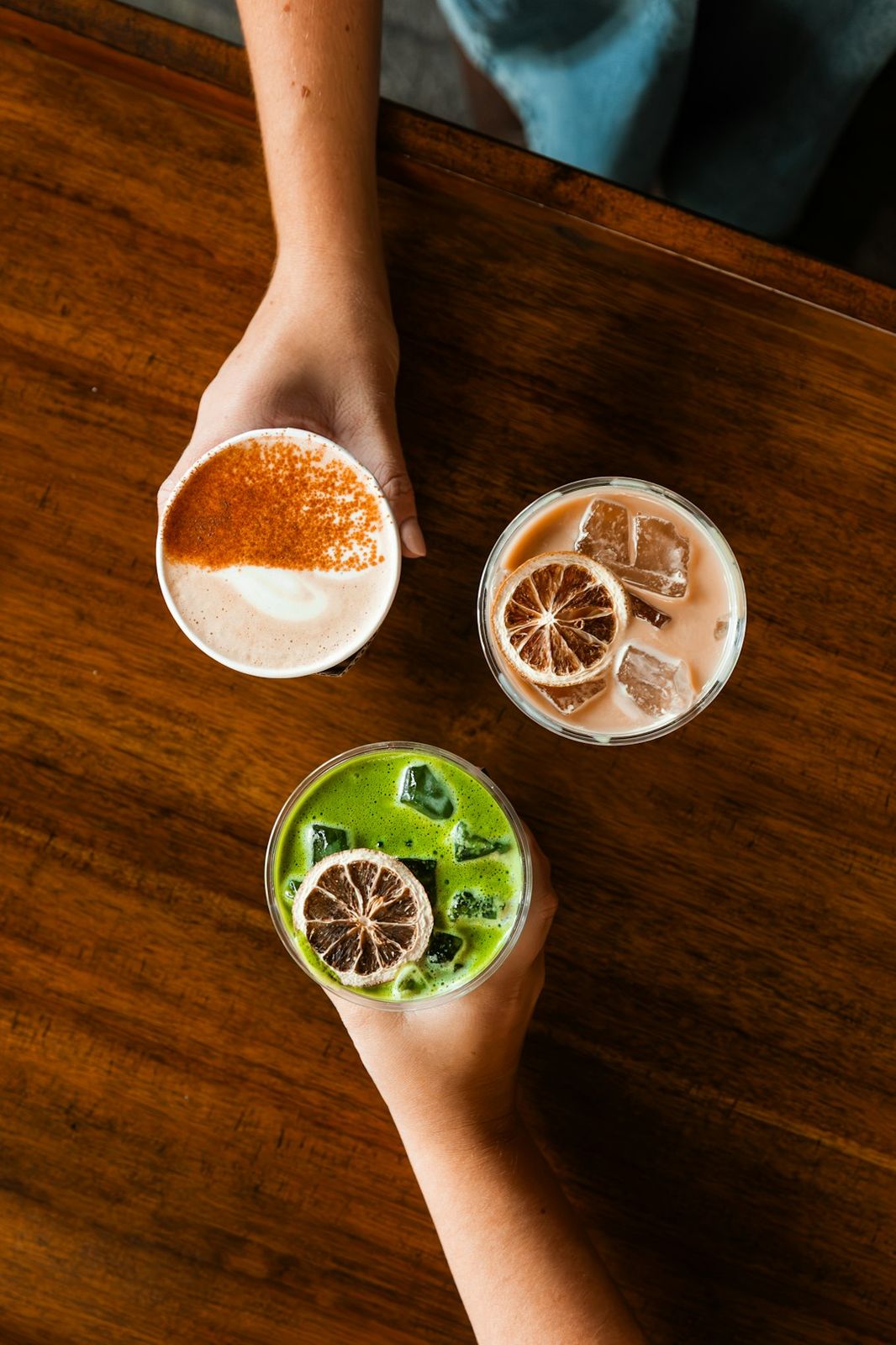 Hands holding three iced drinks with citrus slices on a wooden table