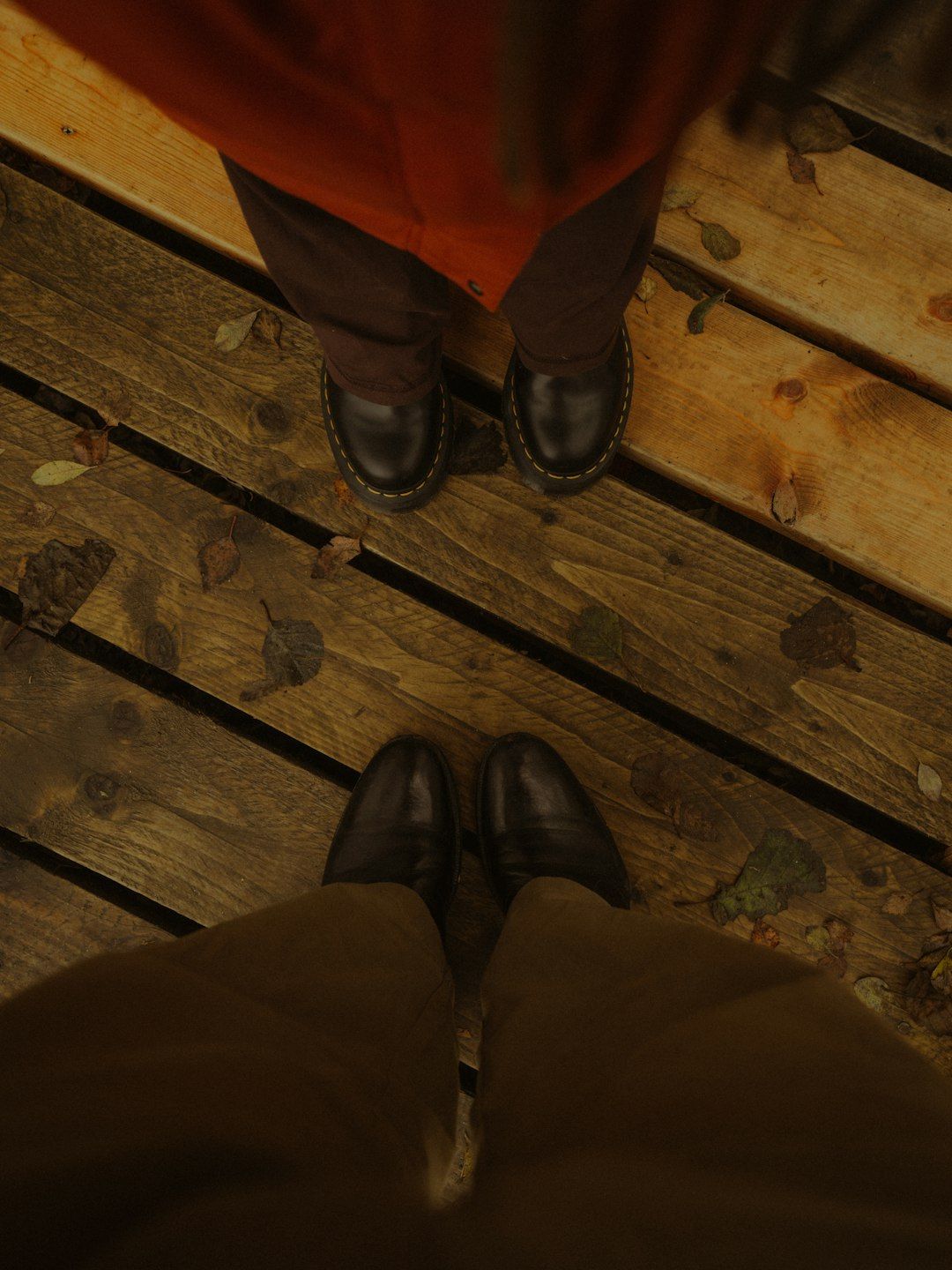 Two people in dark trousers and leather boots standing on a wooden boardwalk with fallen leaves