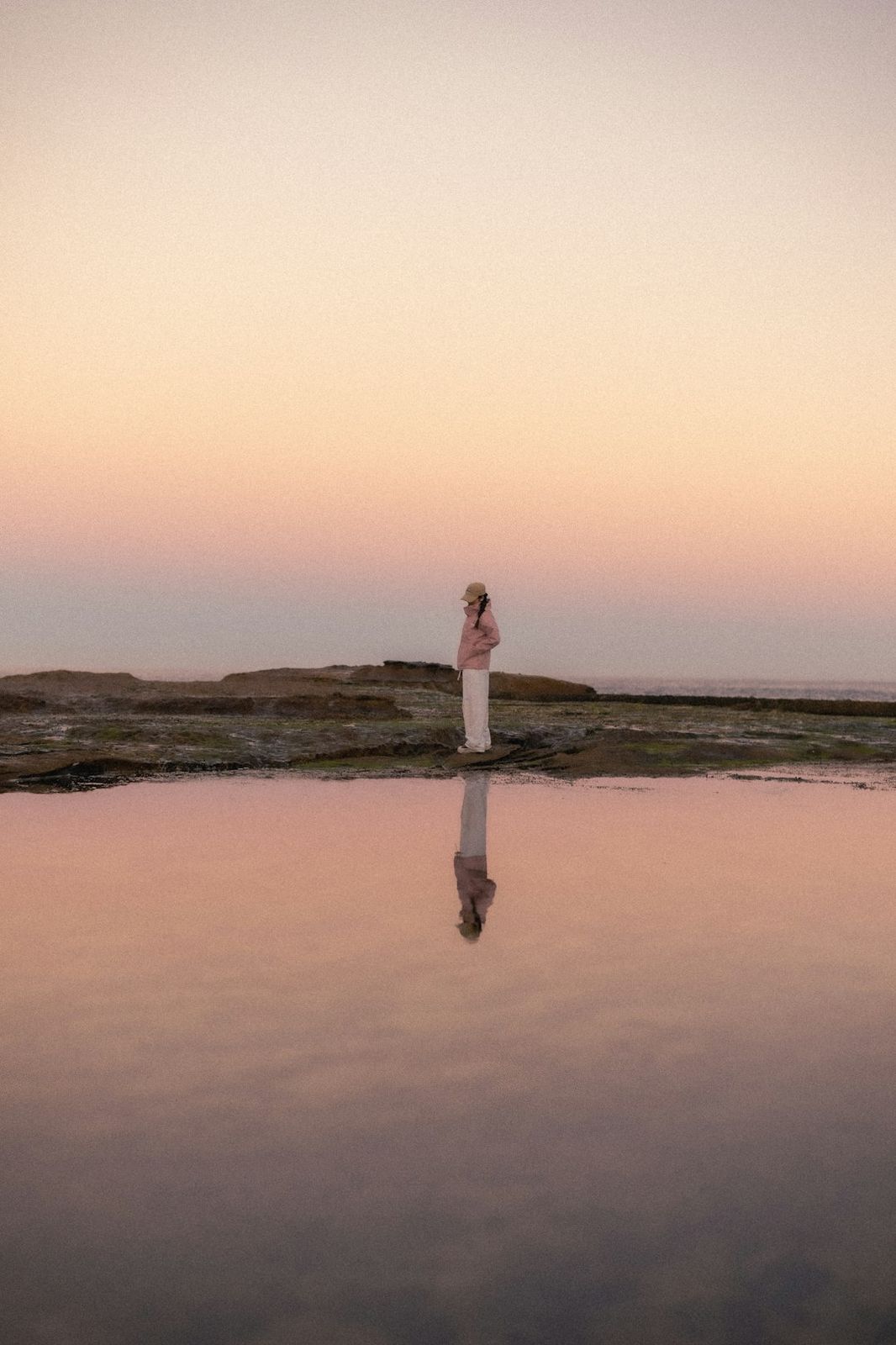A person in a pink jacket and cap reflected in a calm tide pool